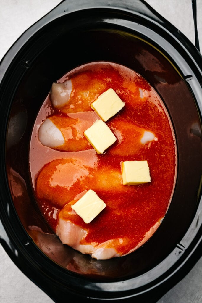 Boneless skinless chicken breasts in an even layer in a crockpot, topped with buffalo sauce and chunks of butter.
