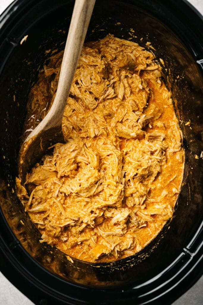Shredded buffalo chicken in a slow cooker with a wood spoon.