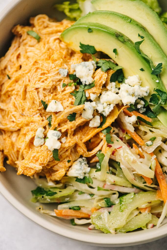 Shredded buffalo chicken in a bowl with coleslaw, avocado slices, and blue cheese crumbles.