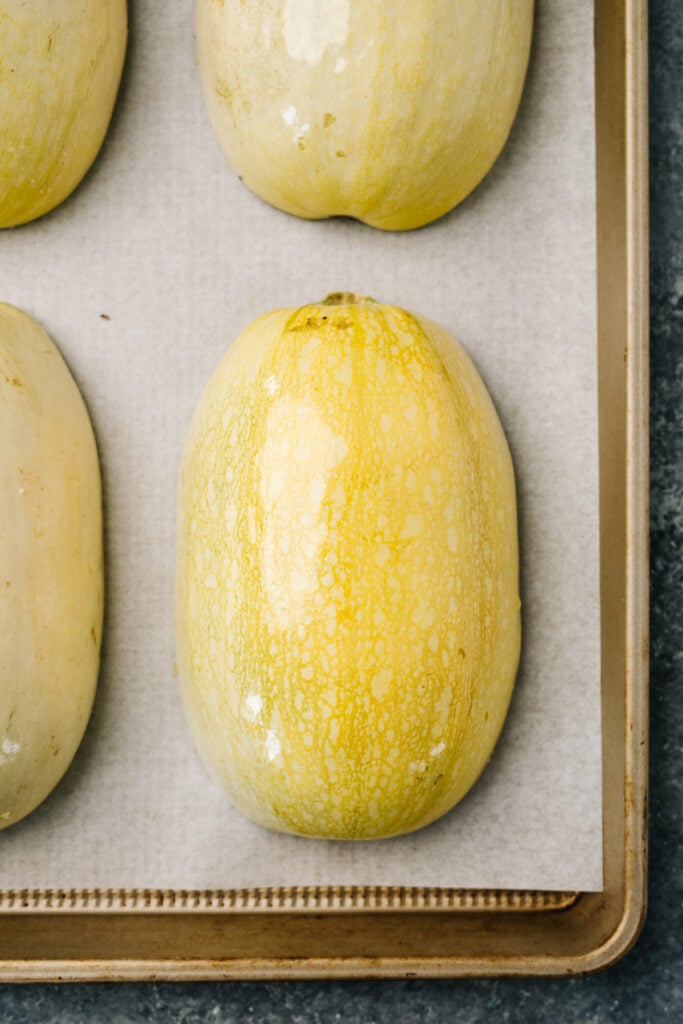 Spaghetti squash halves cut side down on a parchment lined baking sheet.