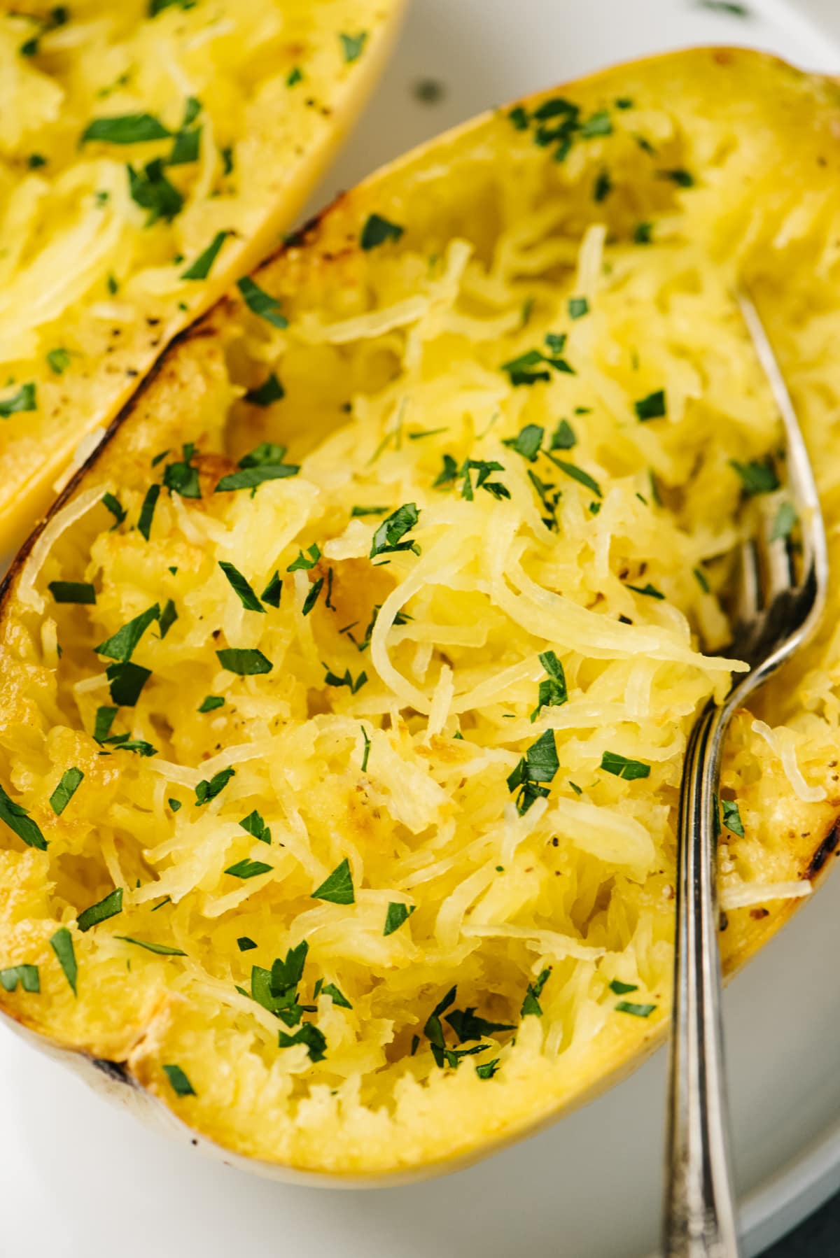 Side view, a fork tucked into roasted spaghetti squash halves, garnished with chopped herbs.