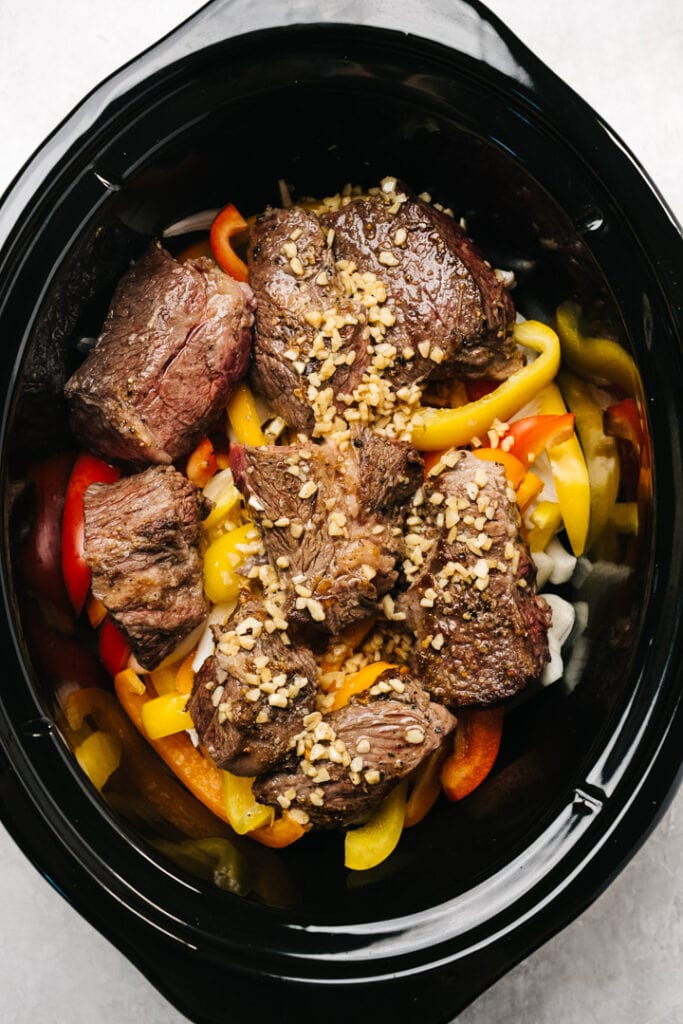 Seared chuck roast pieces over raw onions and peppers in a slow cooker.