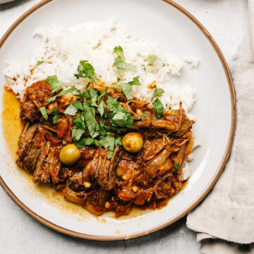 Slow cooker ropa vieja over white rice with lots of fresh cilantro on a concrete background, with a tan linen napkin.