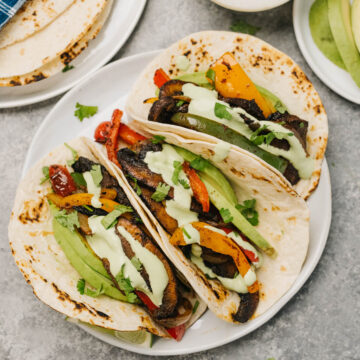 Three vegetarian fajitas on a white plate, stuffed with portobello mushrooms, onions, peppers, and avocado slices, garnished with lime wedges and cilantro lime crema.