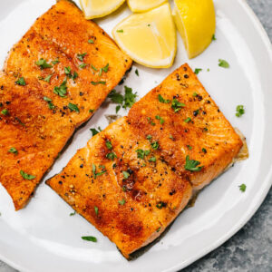 Two pieces of air fryer salmon on a white plate with lemon wedges and chopped parsley.