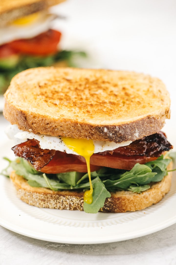 Two breakfast BLT sandwiches on white plates with an egg yolk running down the front of one sandwich.