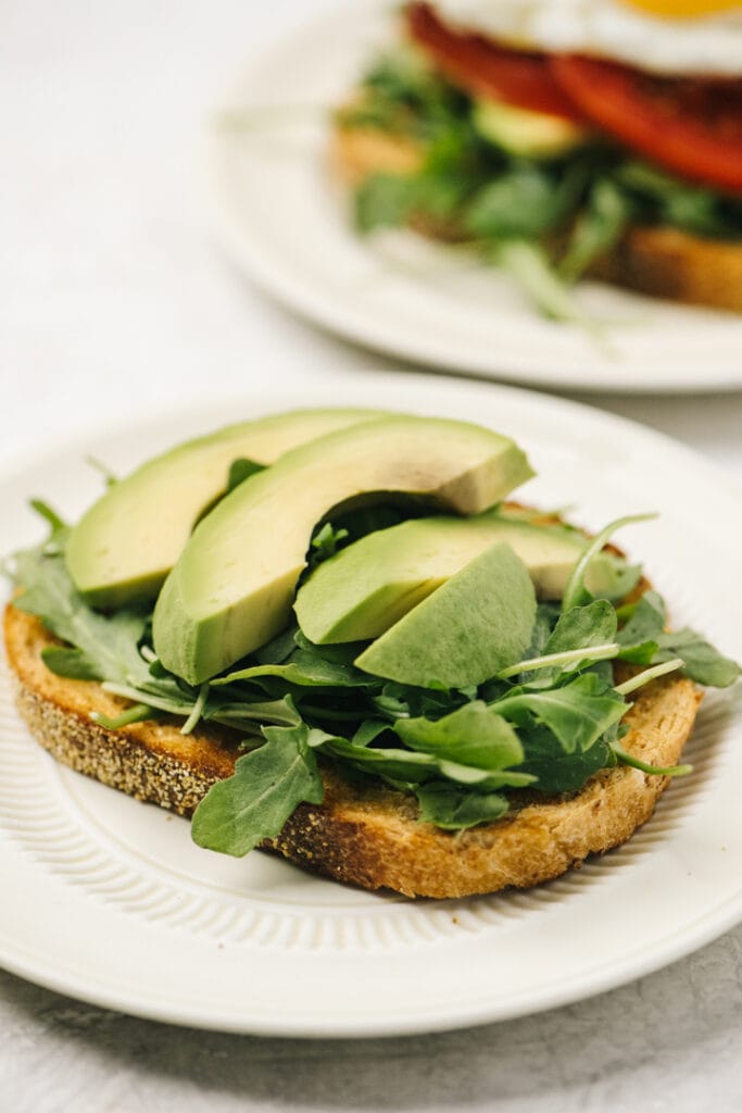 A toasted piece of bread on a white plate topped with arugula and avocado slices.
