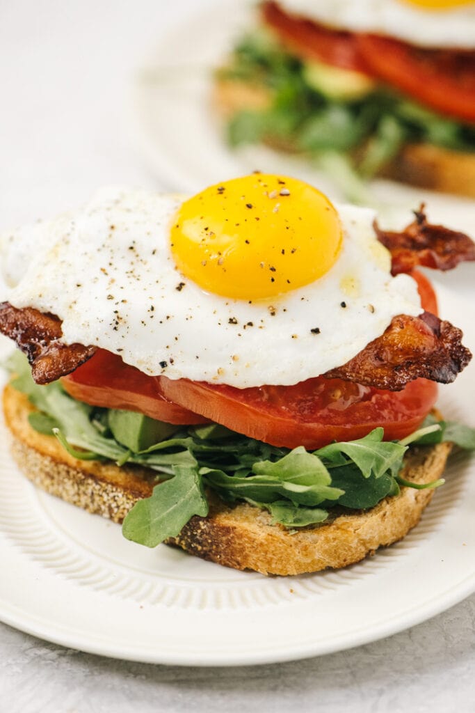 An open face BLT breakfast sandwich topped with a fried egg on a white plate.