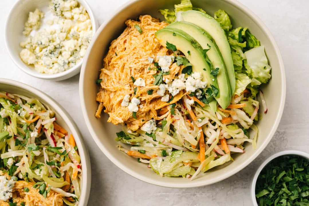 Shredded buffalo chicken, tangy coleslaw, and chopped romaine in a tan bowl, garnished with avocado slices and blue cheese crumbles.