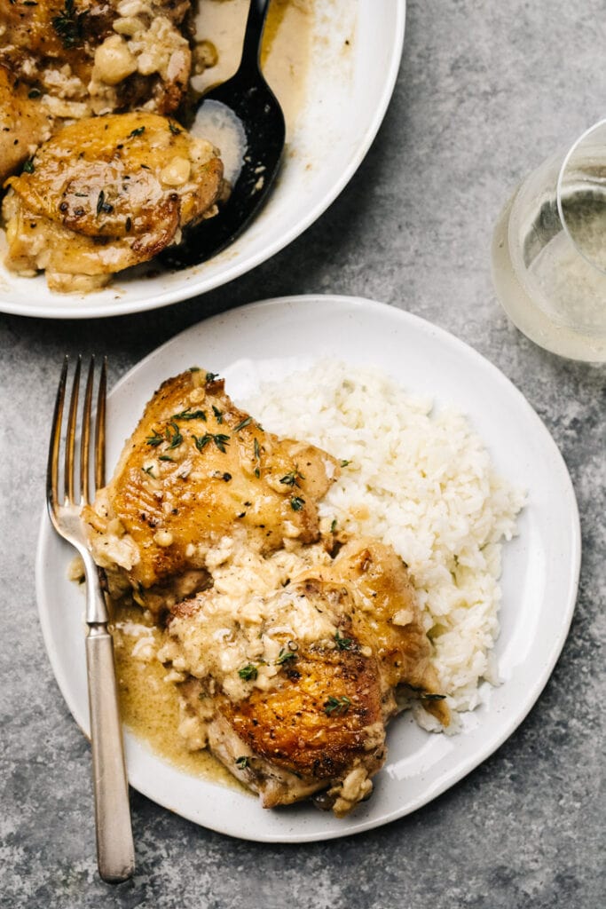 Creamy garlic chicken thighs over rice on a concrete background, with a vintage fork and glass of white wine to the side.