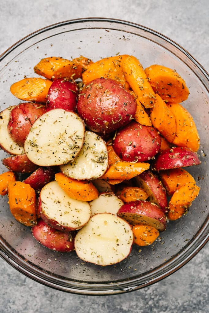 Halved new potatoes and chopped carrots tossed with olive oil, dried herbs, and seasonings in a large glass mixing bowl.