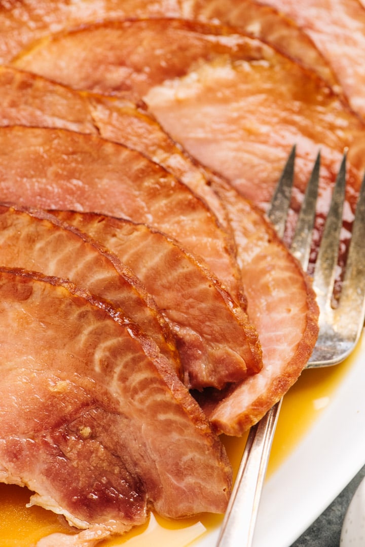 Side view, slices of instant pot ham on a white serving dish with a silver serving fork, drizzled with maple orange glaze
