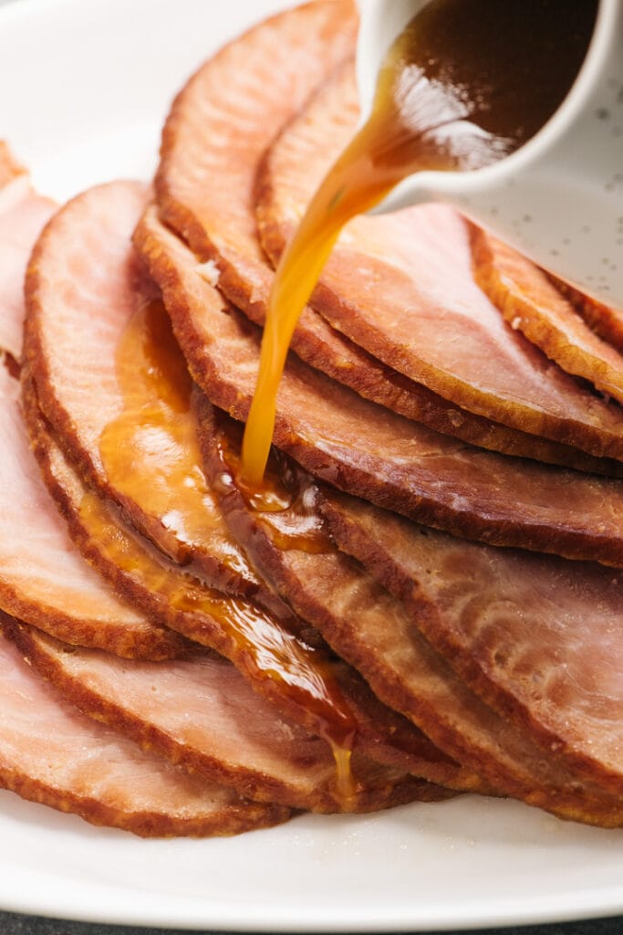 Side view, pouring maple orange glaze over ham slices on a white serving platter.