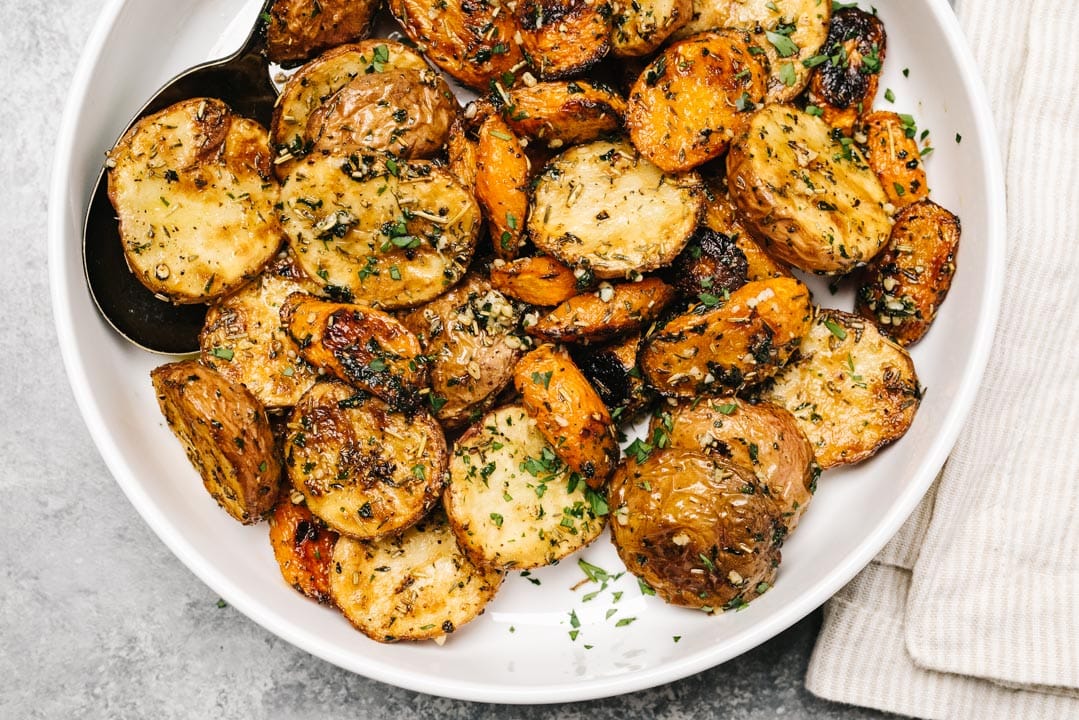 Roasted potatoes and carrots in a white serving bowl on a cement background with a striped linen to the side.