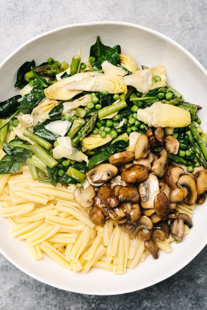 Cooked pasta in a large white bowl topped with sauteed mushrooms, asparagus, artichoke hearts, peas, and spinach.