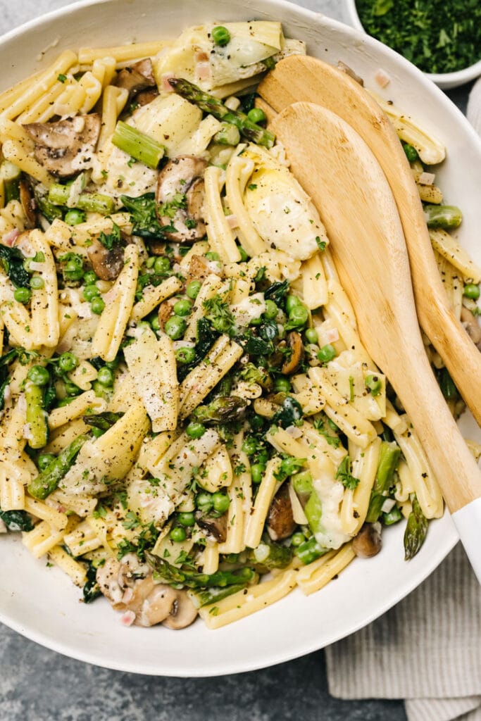 Spring pasta primavera in a large white pasta bowl with wood serving spoons.