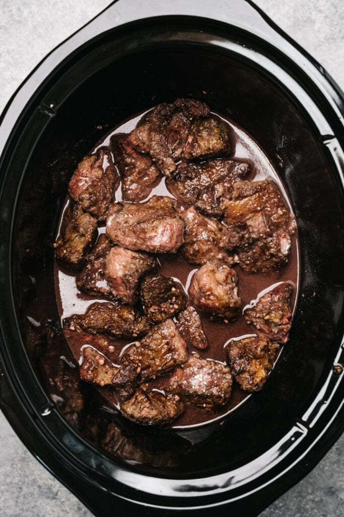 Seared chuck roast pieces in a crockpot, covered with red wine deglazing liquid.