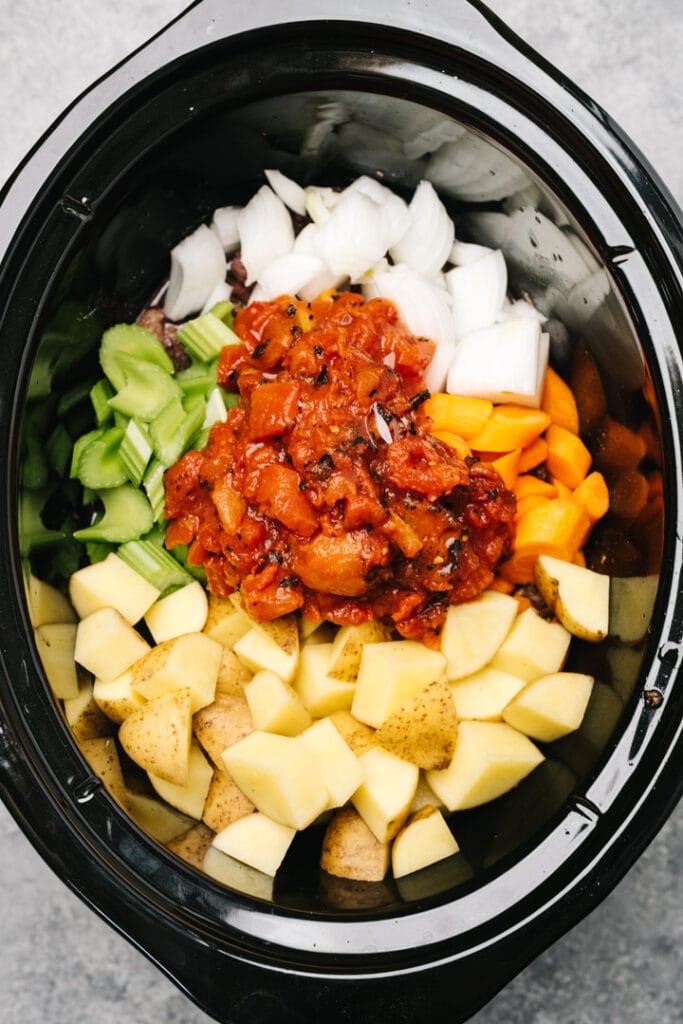 Chopped onions, carrots, celery, potatoes, and canned tomatoes over seared chuck roast pieces in a crockpot.
