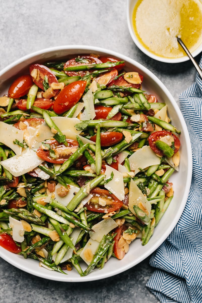 Raw asparagus salad with tomato, parmesan, and pancetta in a white serving bowl with a blue linen napkin and bowl of lemon vinaigrette to the side.