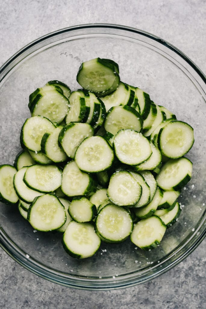 Thinly sliced english cucumbers tossed with salt in a glass mixing bowl.