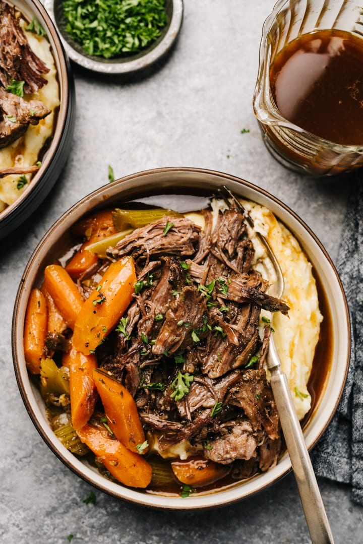 Slow cooker pot roast and carrots over mashed potatoes in a low bowl with a pitcher of gravy on the side.