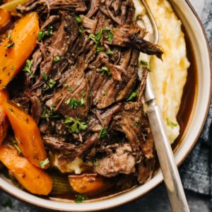 Slow cooker pot roast over mashed potatoes with carrots and celery in a low bowl with a vintage fork and dark grey napkin to the side.
