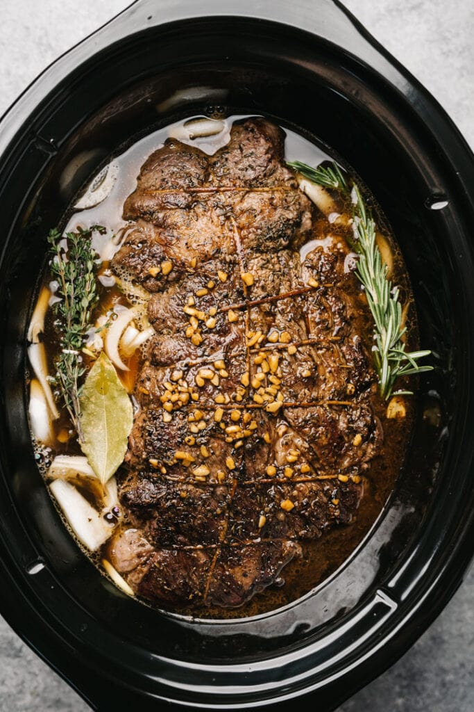 Seared chuck roast over sliced onions in a crockpot with fresh herbs.