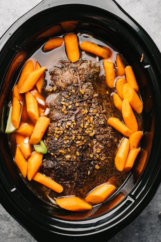 Seared chuck roast in a slow cooker with fresh herbs, carrots, and celery.