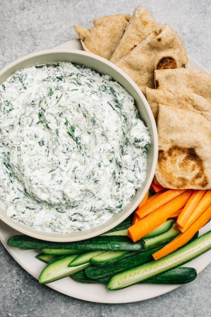 Spinach yogurt dip in a tan bowl on a plate with sliced vegetables and pita bread.
