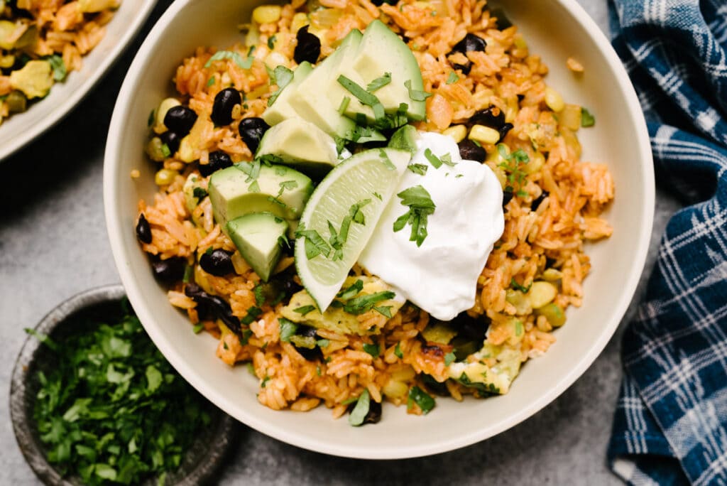 Mexican fried rice in a low tan bowl, garnished with avocado, lime and cilantro, and surrounded by a blue linen napkin and small bowl of fresh chopped cilantro.