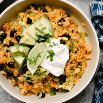 A bowl of mexican fried rice, topped with sour cream, avocado, lime wedges, and fresh cilantro with a blue linen napkin to the side.