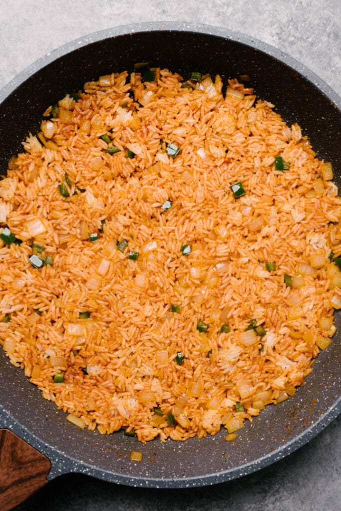 Cooked rice tossed with onion, jalapeno, tomato paste, and seasonings frying in an even layer in a non-stick skillet.