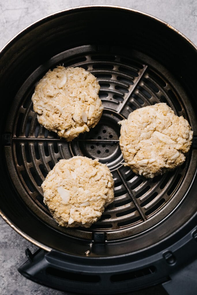 3 uncooked crab cakes in an air fryer basket. 