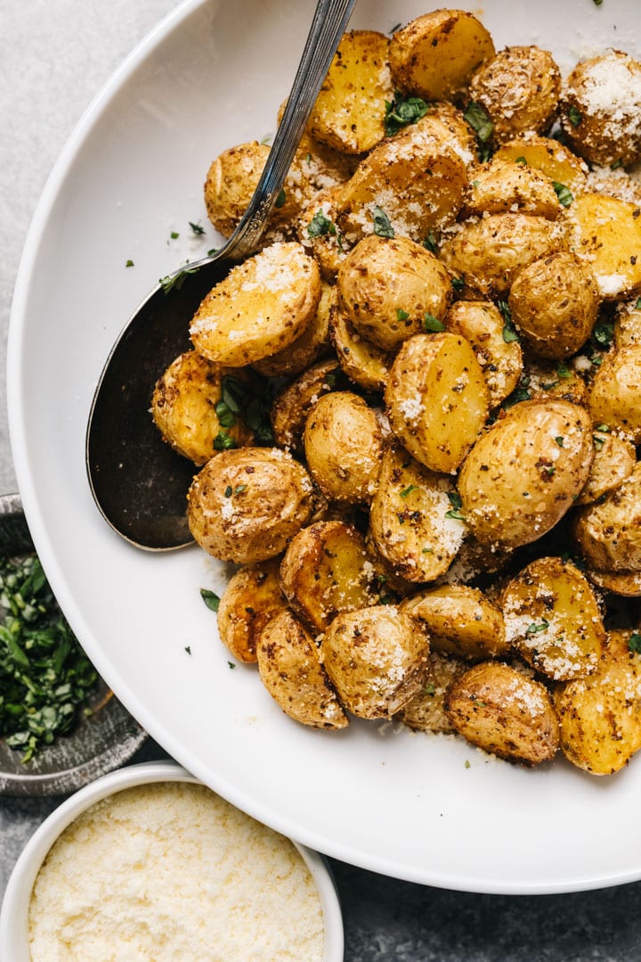 Air fried potatoes in a white serving bowl with a vintage silver serving spoon; chopped herbs and grated parmesan cheese to the side in small bowls.