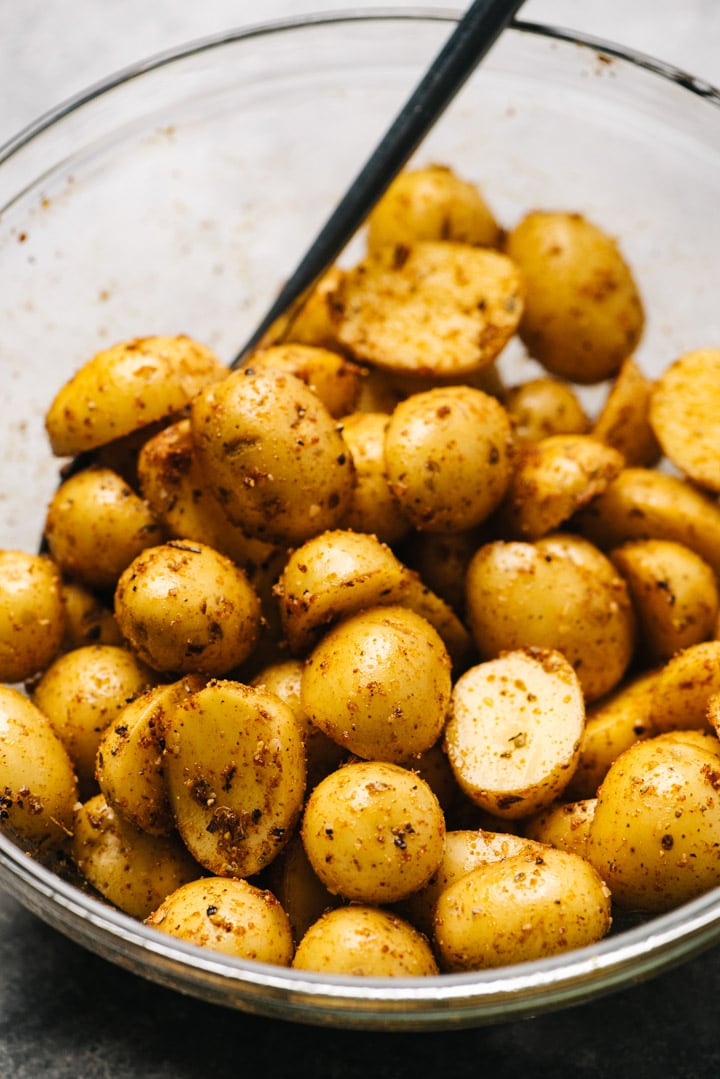 Halved baby potatoes tossed with olive oil and seasonings in a glass mixing bowl.