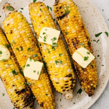 Four ears of grilled corn on a speckled plate, topped with butter and fresh herbs.