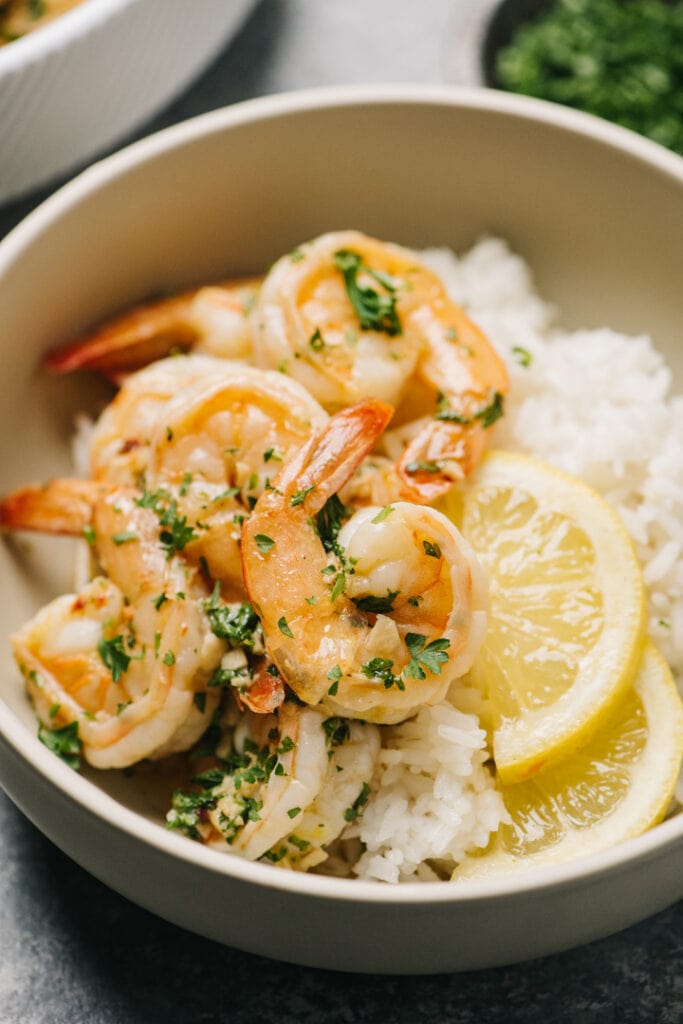 Garlic butter shrimp over white rice in a tan bowl with lemon slices.