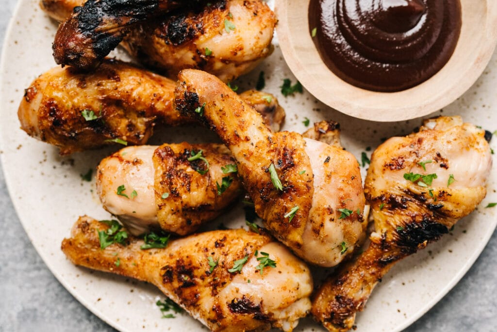 Grilled chicken drumsticks on a speckled tan plate with a small bowl of BBQ sauce for dipping.