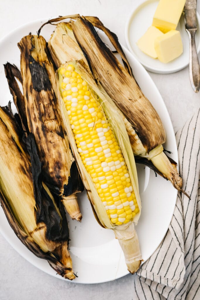 Four ears of grilled corn on the cob with the husks slightly peeled back on a white serving platter with butter and a linen napkin to the side.