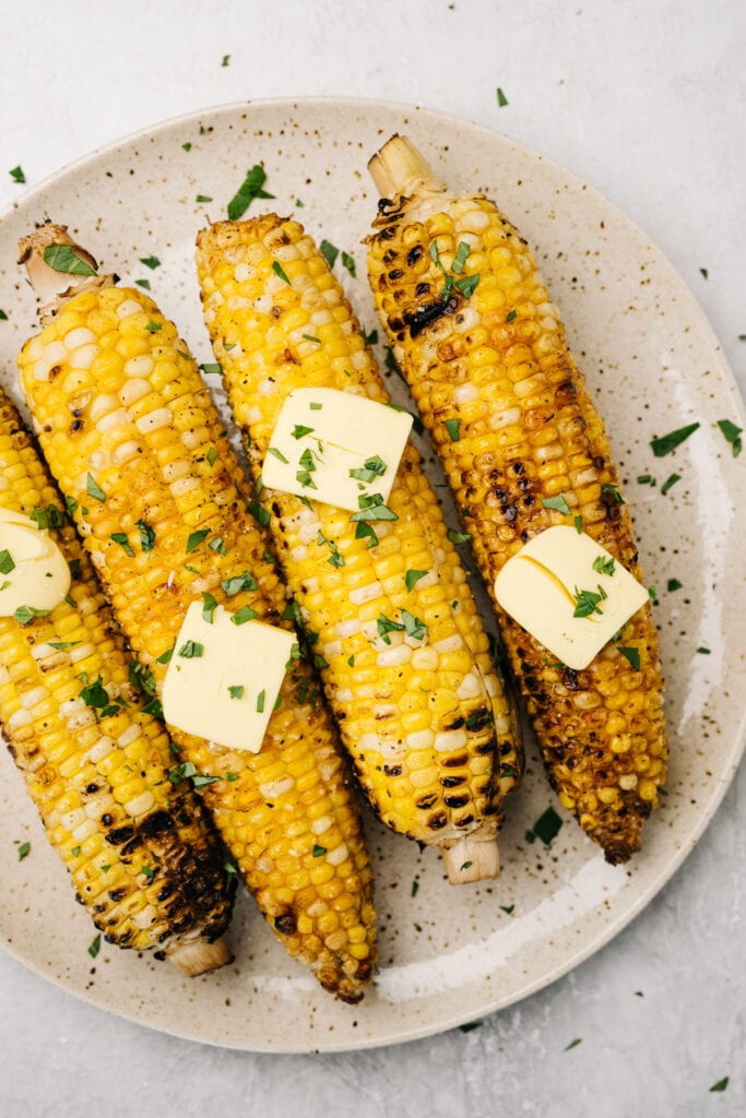 Four ears of grilled corn on a speckled plate, topped with butter and fresh herbs.