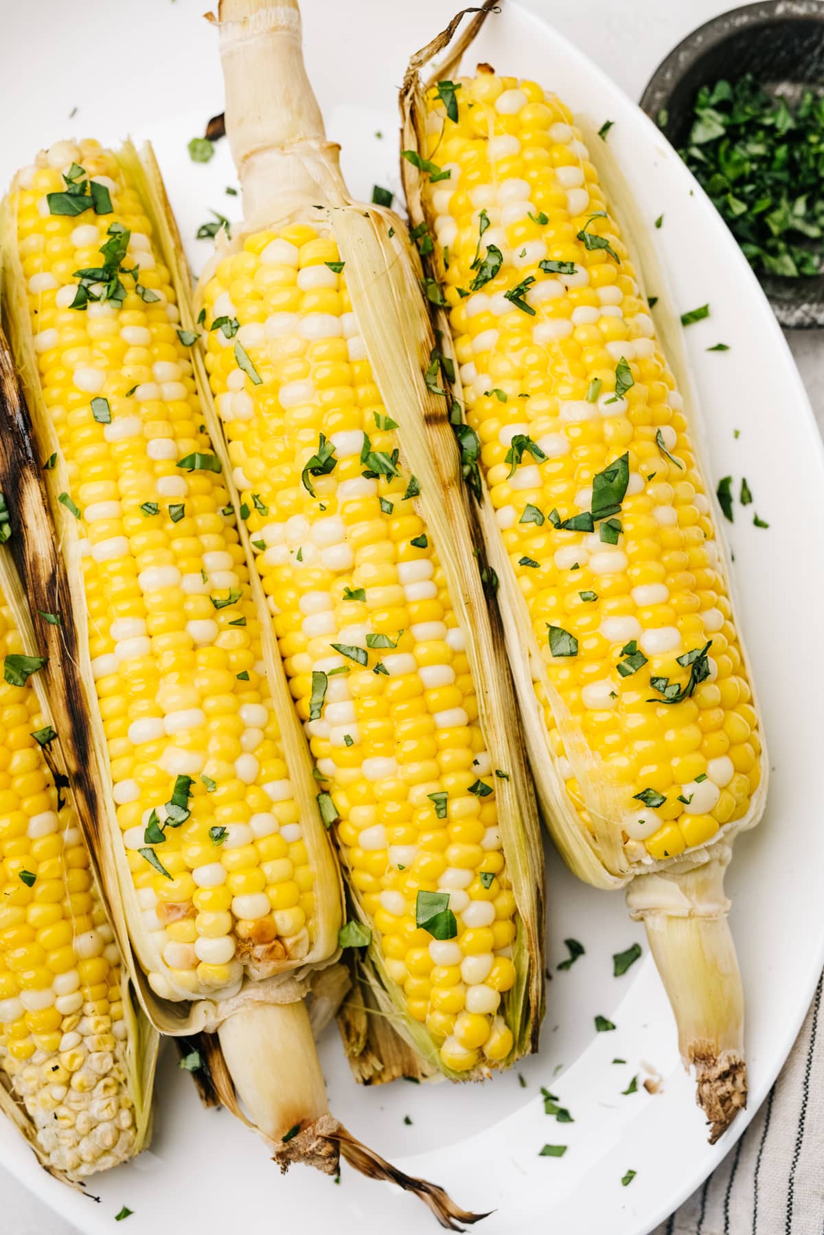 Four ears of grilled corn on the cob on a white plate, with a small bowl of fresh chopped herbs to the side.