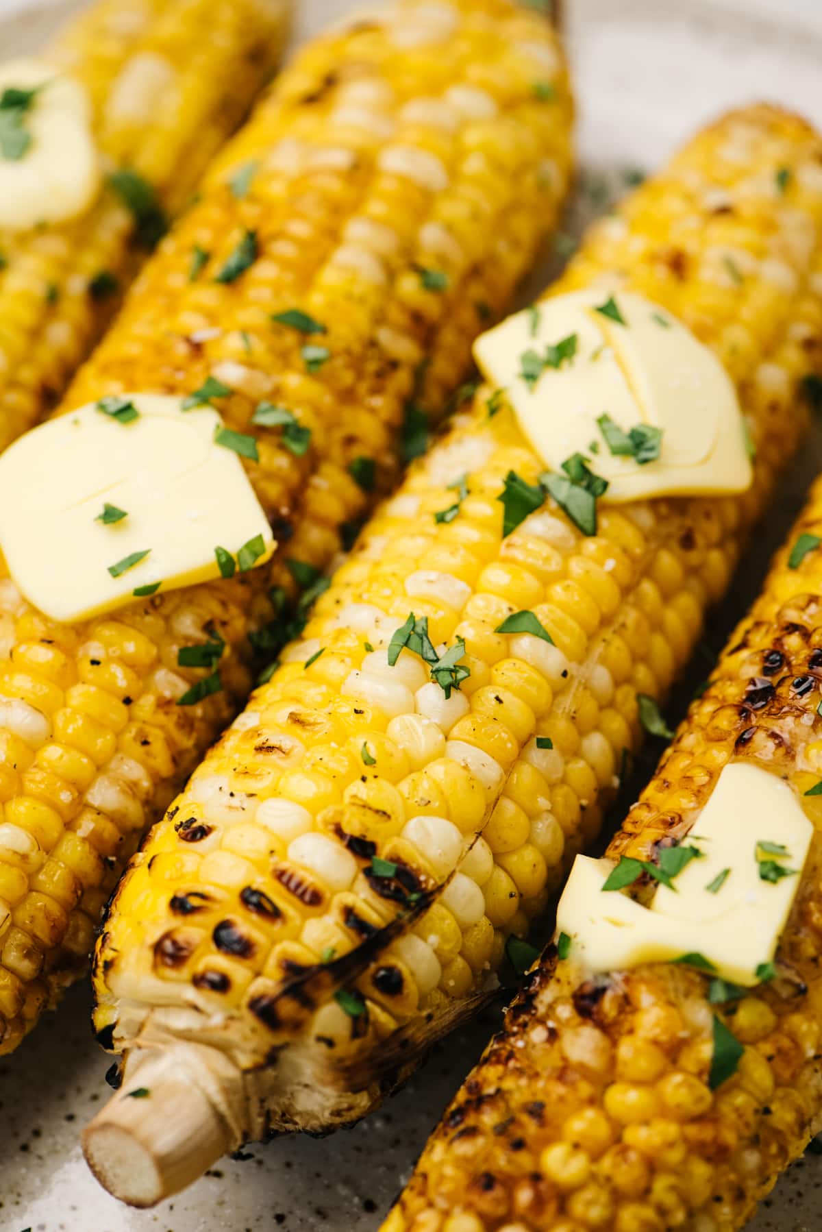 Side view, four ears of grilled corn on a plate, each topped with butter and fresh herbs.
