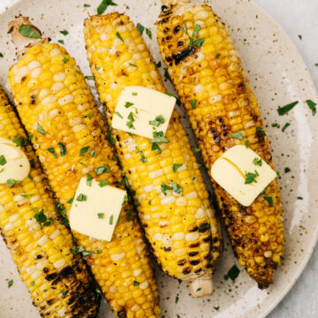Four ears of grilled corn on the cob on a plate, topped with butter and fresh herbs.