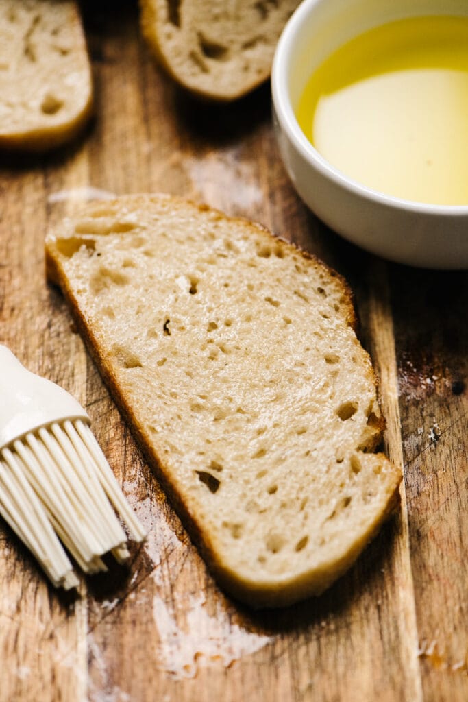 A slice of bread with a bowl of oil and an oil brush. 