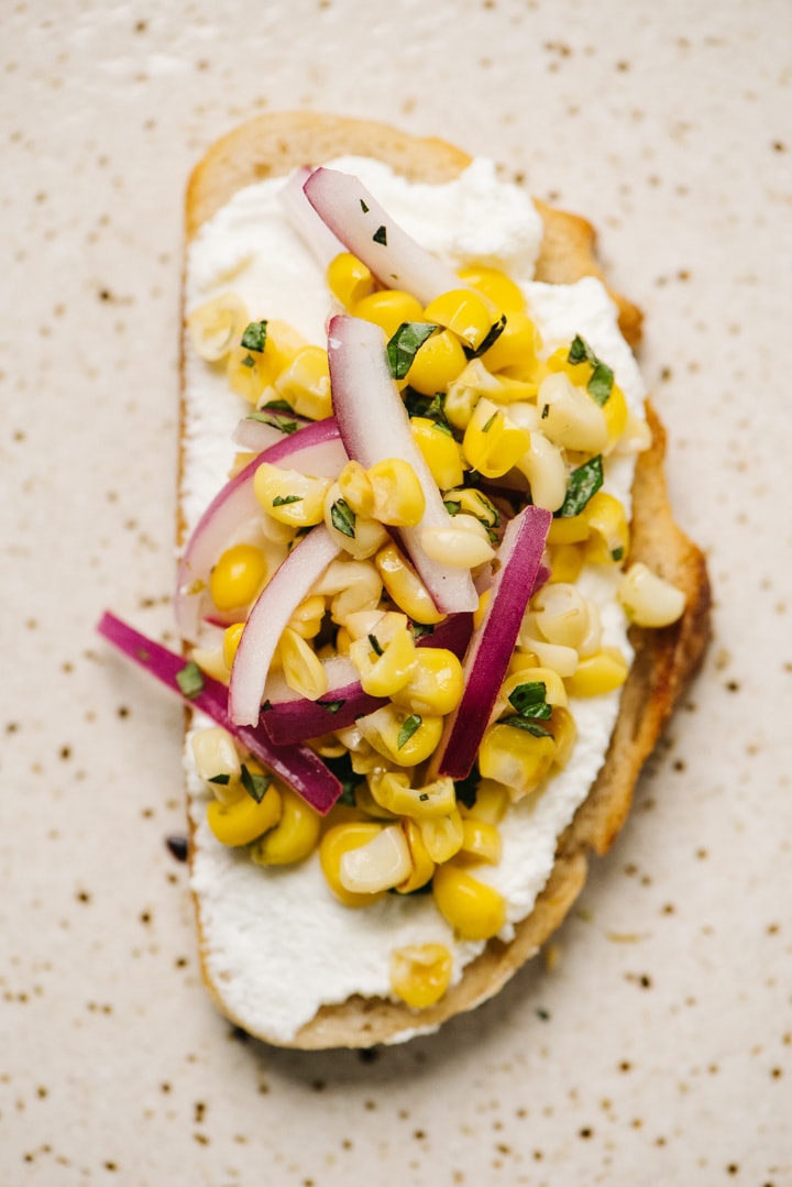 A slice of ricotta toast topped with fresh corn salsa on a speckled tan plate.