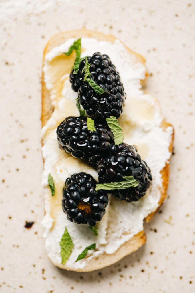 A slice of ricotta toast topped with blackberries, mint, and honey on a speckled tan plate.