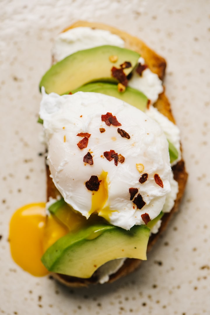 A slice of savory ricotta toast topped with avocado, a poached egg, and red pepper flakes on a speckled tan plate.