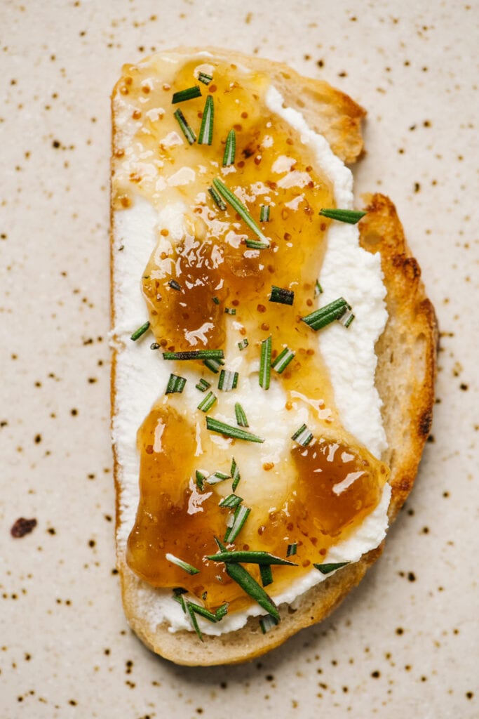A slice of ricotta toast topped with fig jam and fresh rosemary on a speckled tan plate.