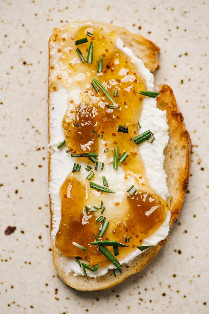 A slice of sweet ricotta toast topped with fig jam and fresh rosemary on a speckled tan plate.