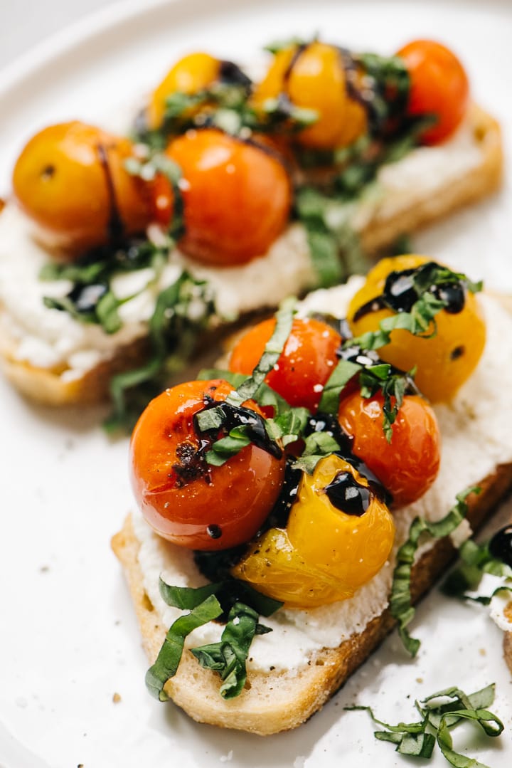 Side view, two slices of ricotta toast topped with burst tomatoes, basil, and balsamic glaze.